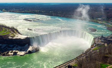 An aerial view of Niagara Falls shows the powerful water cascading over the cliff, surrounded by mist and greenish waters, with people gathering below.