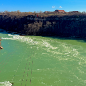 Amidst the rugged cliffs and bare trees, the river flows energetically, with power lines stretching across the view under a clear blue sky.