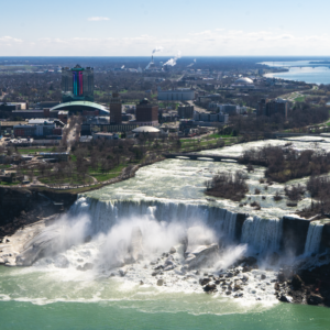 The magnificent Niagara Falls flows powerfully, with the city visible behind and the tranquil river extending beyond the falls.