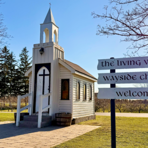 Nestled beneath a sunny sky, the charming Wayside Chapel, marked by a cross and bell tower, greets visitors with a warm welcome sign.
