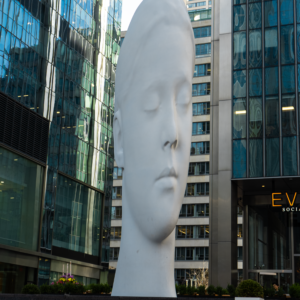 The oversized, smooth white head sculpture, with its peaceful face, creates a striking focal point amidst the modern glass towers of the city.