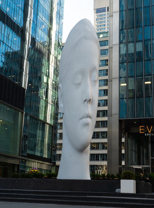 The oversized, smooth white head sculpture, with its peaceful face, creates a striking focal point amidst the modern glass towers of the city.