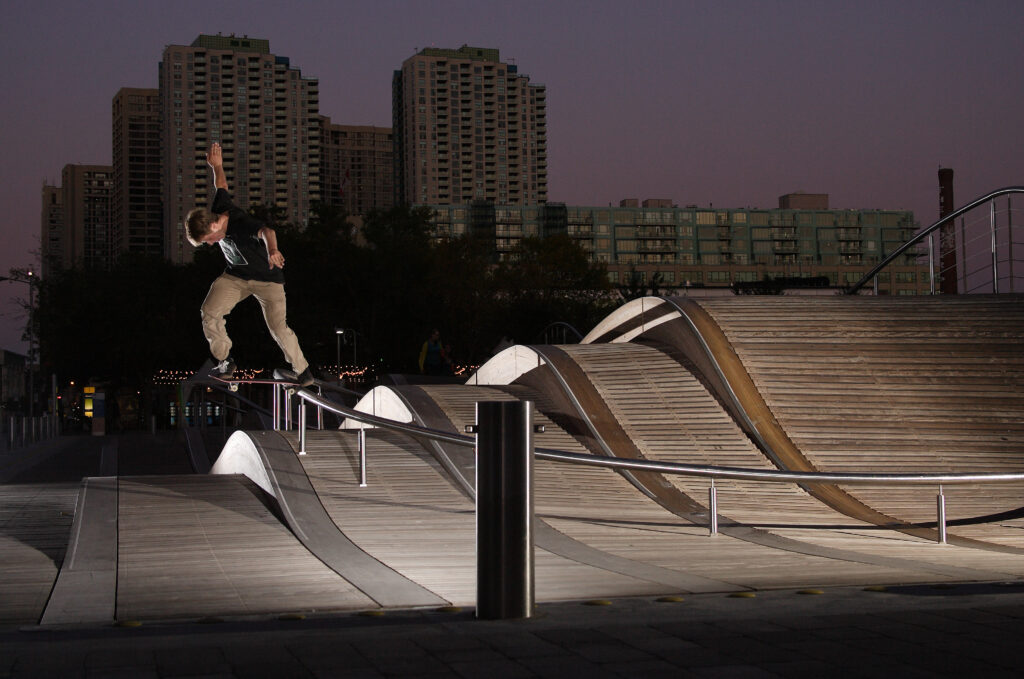 A man performs a skateboard trick on a rail at dusk, surrounded by a vibrant sky transitioning to night.