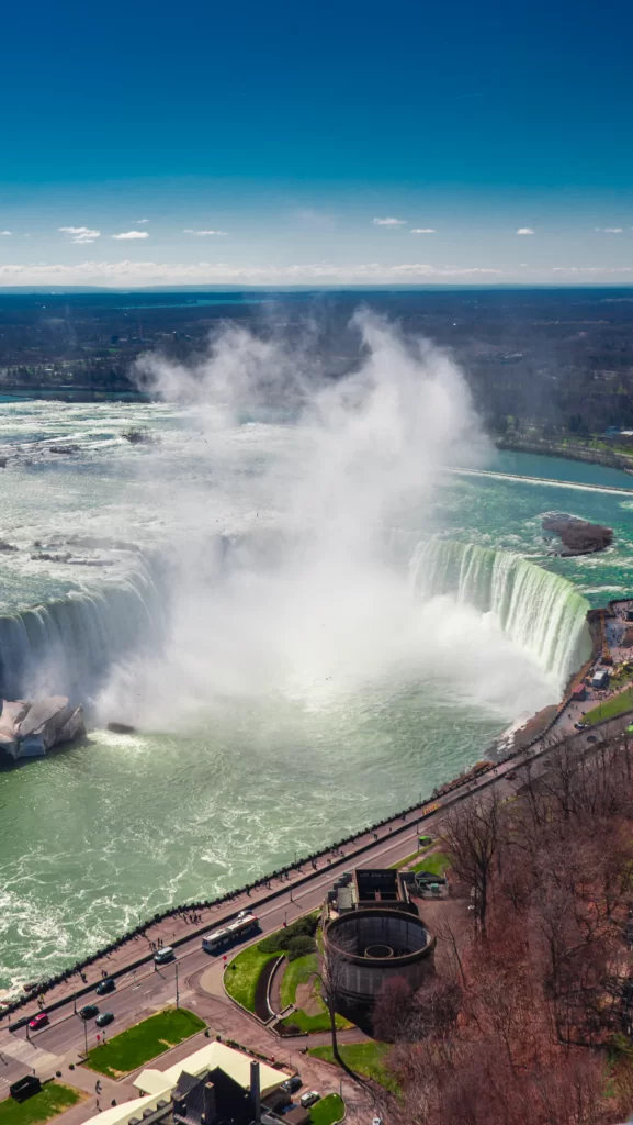 An aerial view of Niagara Falls shows the powerful water cascading over the cliff, surrounded by mist and greenish waters, with people gathering below.