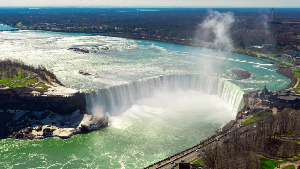 An aerial view of Niagara Falls shows the powerful water cascading over the cliff, surrounded by mist and greenish waters, with people gathering below.