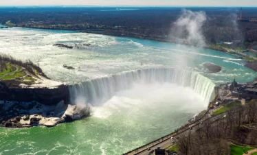 An aerial view of Niagara Falls shows the powerful water cascading over the cliff, surrounded by mist and greenish waters, with people gathering below.